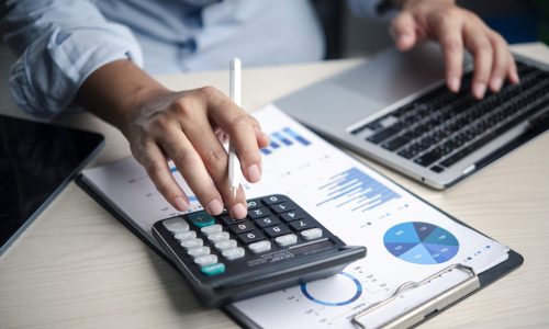 Calculator, finance Analyst presses calculator on budget report with charts laptop in office, reviewing data, accounting, numbers, analysis, planning, costs, and profit for decisions.
