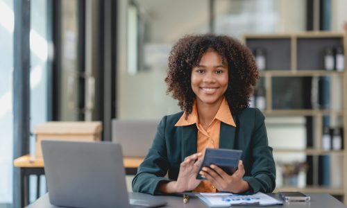 Young african american business woman holding calculator and working with computer at office desk.