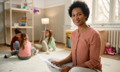 Smiling African American preschool teacher sitting on the floor and looking at camera. There are kids playing in the background.