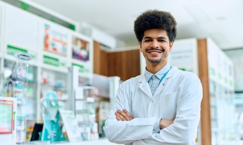 Happy pharmacist standing with arms crossed while working in a pharmacy and looking at camera.