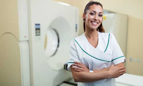 Happy female doctor with arms crossed standing next to MRI scanner and looking at camera.
