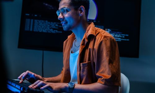 A serious young cyber threat analyst holds a pen while thinking at the desk in a blue-lit cyber operations centre. The frame illustrates defending the corporate network.