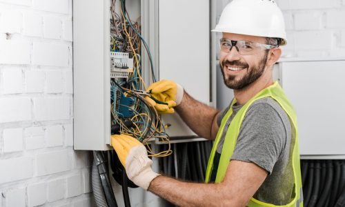 smiling handsome electrician repairing electrical box with pliers in corridor and looking at camera