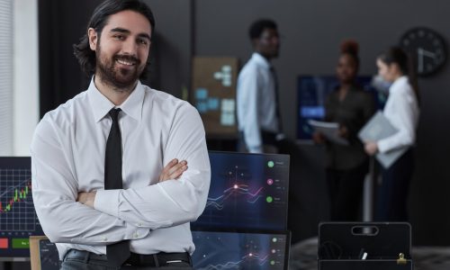 Portrait of professional young trader smiling at camera while standing with his arms crossed in office