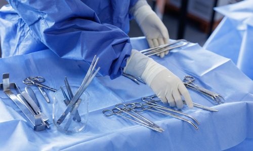 Gloved clinician organizes stainless surgical instruments on a blue sterile field, showing OR preparation and infection-control workflow.