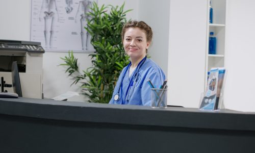 Portrait of medical assistant working at hospital reception desk, using computer with healthcare appointments and report papers. Nurse doing registration work with checkup visit forms in clinic lobby.