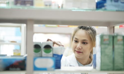 Female smart young-adult pharmacist in professional white gown looking and checking stock inventory in modern pharmacy, drugstore indoor holding clipboard. Pharmacy, medicine, and healthcare concept.