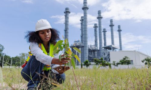 A woman in safety gear and a hard hat plants a small tree near an industrial facility. The image reflects eco-awareness, sustainability, environmental responsibility, and the balance between industry and nature.