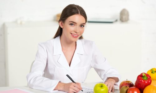 smiling dietitian in white coat writing at workplace with fruits and vegetables on table