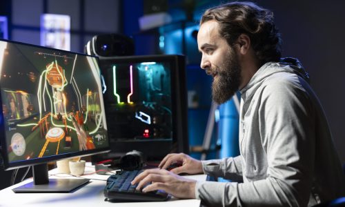Joyous man in dark living room playing science fiction videogame on gaming PC, concentrating on shooting foes. Radiant gamer battling flying robots with futuristic gun in online singleplayer shooter