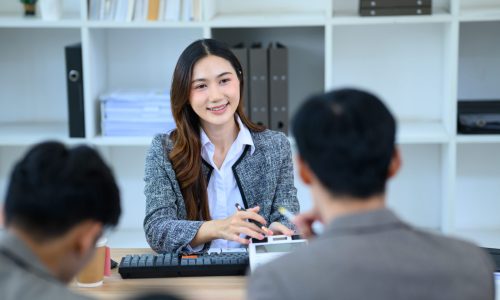 A confident young Asian female advisor smiles warmly while meeting with two clients at her desk, ideal for finance, banking, and business consulting concepts.