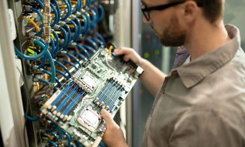 Close-up of serious bearded IT support specialist standing by cabinet of mainframe and examining motherboard of server